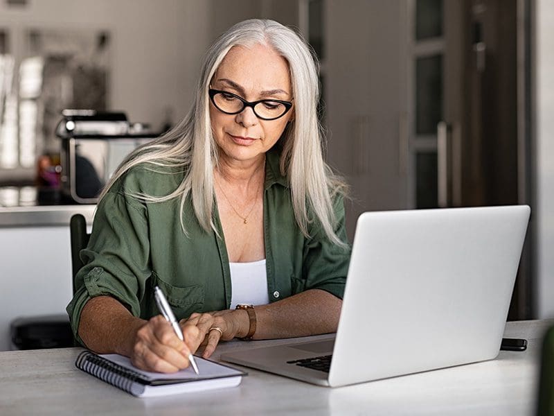 Woman writing in a notebook with her her laptop at a table.